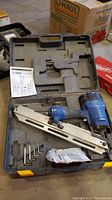 Campbell Hausfeld blue air framing nailer inside open carrying case with instruction manual and hex keys. The tool shows surface wear and is placed in a molded plastic case with yellow latches.