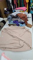 Top view showing a folded light beige wrap cardigan sweater in the foreground with a stack of various tops in different colors and patterns behind it.
