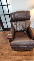 Front view of brown leather recliner chair with padded headrest and armrests, remote control on seat.