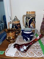 Photo of German beer steins, porcelain creamers, and glass bird paperweight arranged on a table showing detailed designs.