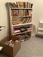 White bookshelf filled with books on four shelves and a cardboard box on floor with books inside.