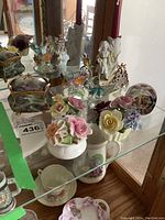 Photo showing glass birds, ceramic floral sculptures, small trinket box with lid, and candle holder on a glass shelf in front of a mirror