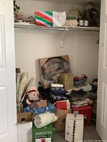 Closet photo showing many Christmas decorations including wrapping paper rolls, gift bags, wreath box, and assorted holiday items piled on shelves.