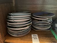 Two stacks of mostly white ceramic collector plates with various colorful designs and hanging hardware, placed on wooden shelf.