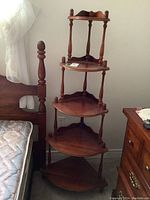 Full view of solid wood corner shelf positioned between bed and dresser showing all five shelves and turned spindle supports.
