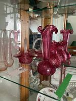 Photo showing deep cranberry red glassware: basket with handle and ruffled edge, pitcher with bulbous base and flared opening, and a pair of bud vases with narrow necks and flared rims.