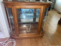 Front view of the wooden display cabinet showing the glass door with a metal handle, adjustable glass shelf inside, and curved glass side panels.