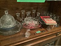 Display of all glassware items on wooden shelf, including candle holders, butter dish, bowls, vase, and spreaders. Some candle holders tied with pink ribbon.