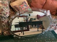 Full view of oval wall mirror resting against a couch with floral cushions, showing the etched flower pattern on the glass frame and clear reflective mirror surface.