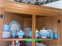 Dinnerware including plates, cups, saucers, teapot, cream and sugar sets arranged inside a wooden cabinet with glass doors.