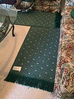 Photo of one green runner with pink and white floral pattern, fringed edges visible, placed along a sofa and carpeted floor.