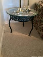 View of one metal framed glass top side table showing round glass surface and ornate metal apron with curved legs.