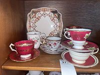 Four vintage teacups and their matching saucers displayed on a wooden shelf alongside a decorative serving plate with floral designs.