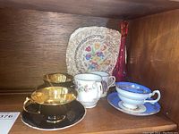 Two Paragon black and gold teacups on matching saucers with additional white and blue floral teacups and porcelain plate in background