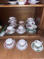 Eleven assorted vintage teacups and saucers displayed on a wooden shelf, featuring floral patterns and gold accents.
