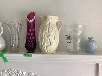 Photo showing a line of vases on a shelf including clear glass fluted vase, purple hobnail glass vase, white ceramic pitcher with relief figures, frosted glass vase, and small blue Spode vase