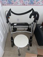 Walker with four wheels, padded handles, black seat, and brake handles captured against a white wall.