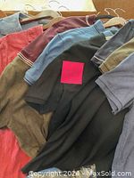Photo showing men's sports shirts hanging on hangers, various colors including black, blue, red, olive green, burgundy. Shirts have collars and short sleeves.