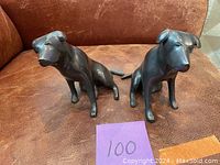 Two metal dog sculptures, dark patina, sitting poses with smooth polished finish on brown leather surface.