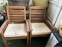 Two wooden patio chairs with white cushions placed side by side. Chairs have slatted backrests and armrests, showing signs of outdoor wear.