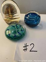Three distinct glass paperweights displayed together on a white surface, showing different colors and shapes.