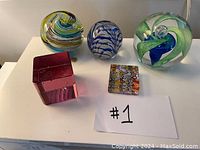 Overview of all five glass paperweights on white surface: three round swirl patterns and two square/rectangular shapes including rose pink cube and multicolored millefiori