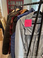 Photo shows nine men's outdoor jackets and vests hanging on a rack, including a blue down vest, shearling-like plaid jacket, leather jackets, lightweight windbreakers, and a puffy coat.