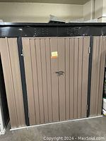 Front view of brown vertical metal panel garden shed with black metal trim around doors and roof edges. Double doors have a locking latch and small yellow note with number '132'.