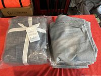 Two sets of grey curtains on a red surface; one folded out and one sealed in plastic with a white ribbon and label showing product details.