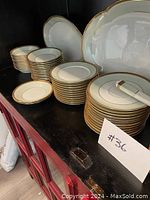 Stacks of plates and bowls in cream with gold trim arranged on a black shelf, showing dinner plates, dessert plates, smaller plates, and bowls with matchng serving tool.