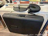 Closed black portable massage table with handle and metal clasps seen from front and top edge.