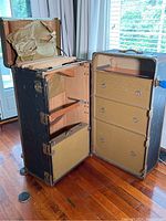 Front view of the open steamer trunk showing three drawers with ring pulls and compartments with tiedown straps inside, lined with aged paper.