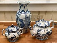 Full view of a blue and white hexagonal teapot, a matching covered jar, and a tall blue and white vase, all positioned on a wooden floor against a beige wall.