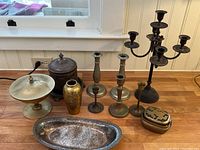 Full view of all items in the lot arranged on a wooden floor near white panel wall beneath window showing the 15-inch silverplate candelabra, candlesticks, brass vase, oval tray, biscuit tin, and lidded container.