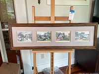 Wide shot of entire framed set with four lithograph prints arranged horizontally inside a single long frame on an easel.