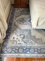 Full view of one corner of the large rectangular rug placed on wood flooring next to a bed and a wooden dresser, showing the intricate geometric and floral iris pattern in multiple blue tones.