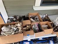 Nine assorted cardboard boxes filled with vintage hardware pieces including door knobs, locks, plates, and wrapped items on a table in front of a house exterior.