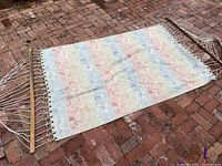 Full view of the hammock laid out on brick surface, showing the faded multicolor striped canvas fabric and attached rope ends with wooden spreader bar.