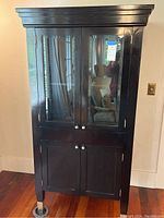 Full front view of the tall black wooden storage cabinet with closed doors showing upper glass panels and lower solid wood doors with silver knobs.