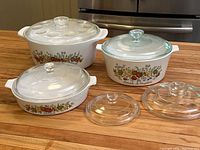 Three CorningWare Spice of Life casseroles and two additional lids displayed on wooden surface in front of stove.