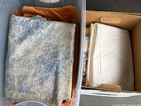 One box shows folded blue and white floral fabric on top with solid orange velvet fabric underneath and a second box with white delicate fabric folded inside.