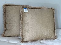 Two large gold/tan throw pillows leaning against a white wall, showing front and back textures.
