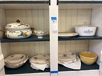 Two wooden shelves displaying an assortment of china and CorningWare including a large covered metal casserole, multiple plates, bowls, and a yellow mixing bowl