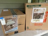 Two large cardboard boxes with printed images showing the two hanging lamps inside, one with milk glass globe and one with floral design globe, packed for storage.