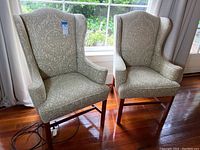 Two vintage wingback chairs positioned side by side near a window, showing overall front view, fabric pattern, and wooden legs.