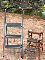 Photo showing both stools side by side on brick patio: rusty vintage Cosco kitchen stool with round top, folding mechanism, and newer metal folding step stool with black tread steps.