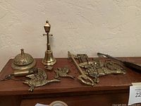 Photo showing all brass items on a wooden surface, including cast brass geese, a decorative bell, brass lamp post figurine, abacus-style piece, shoehorn with key, small lock and key.