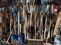 Wide view of various garden tools including shovels, spades, rakes, and brooms grouped together against a wall in a storage shed.