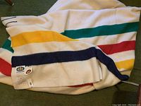 Blanket laid out showing green, red, yellow and navy stripes on white wool, partially folded to display tag
