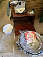 Waring Pro Professional Meat Grinder displayed on wooden bread box alongside a glass baking dish and metal wok with lid wrapped in plastic along with recipe booklet.